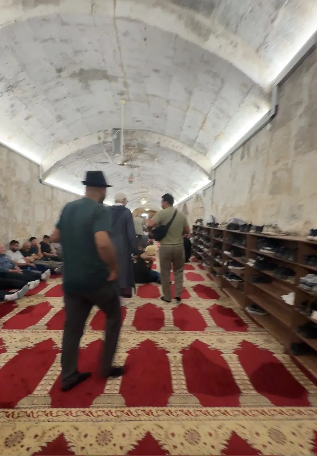 Intérieur voûté en pierre d'une salle de prière avec tapis rouge, visiteurs au repos.
